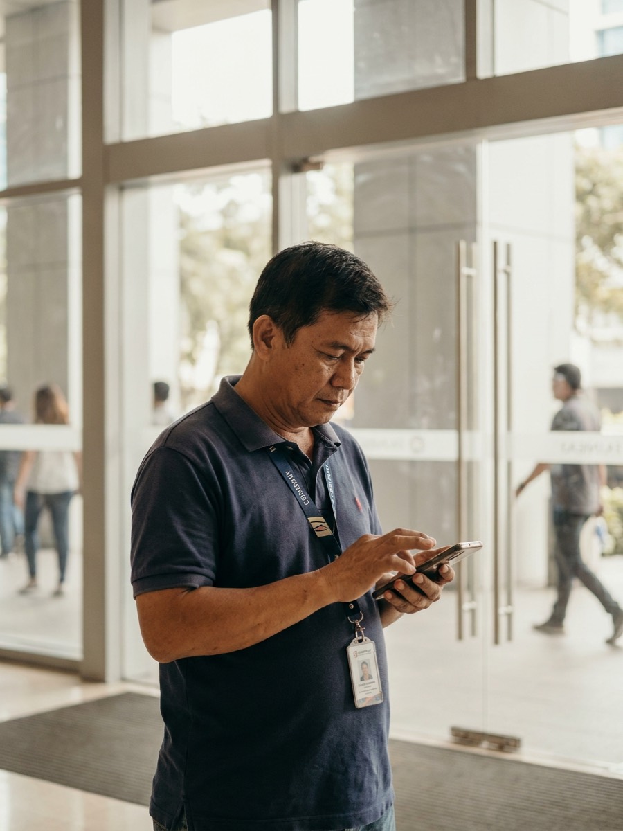 Filipino maintenance worker in a company uniform clocking in on his phone at the lobby of a client office building